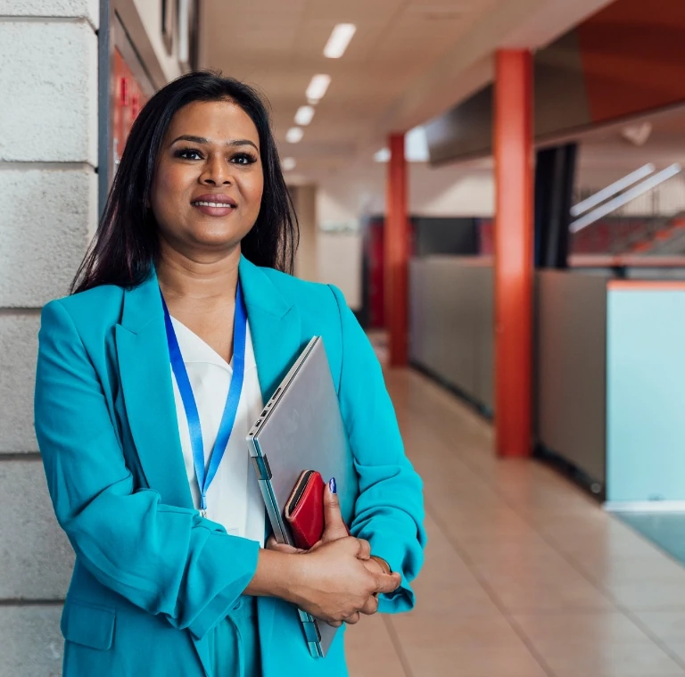 Smiling female teacher standing in a school hallway, carrying a laptop